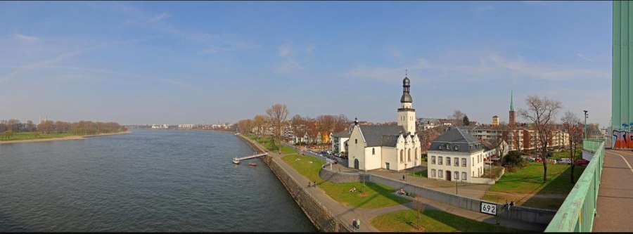 A wide riverside view of Cologne Mülheim, showing the Rhine river, a church with a spire, and buildings along the promenade, seen from a bridge.
