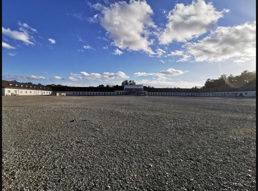 A wide view of the gravel roll call yard at the Dachau Memorial Site, with long low former camp buildings along the horizon under a blue sky with scattered clouds.