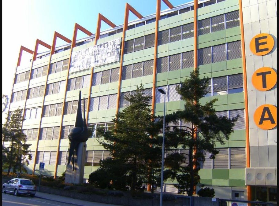 A modern multi story ETA watch factory building in Grenchen, Switzerland, with large orange structural beams and an ETA logo mounted on the facade.