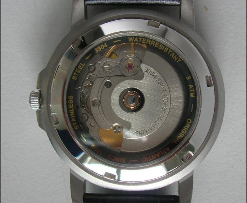 Close up photograph of the back of a wristwatch with its case back removed, showing a metal automatic watch movement with a partial rotor, jewels, and engraved text including Swiss Made and Twenty Five Jewels.