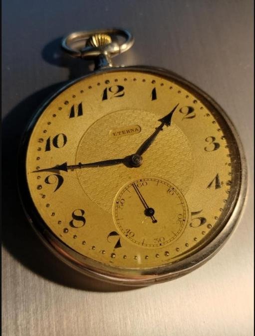 A round silver toned Eterna pocket watch with a gold dial, marked ETERNA above the center, arabic numerals, and a small seconds subdial.