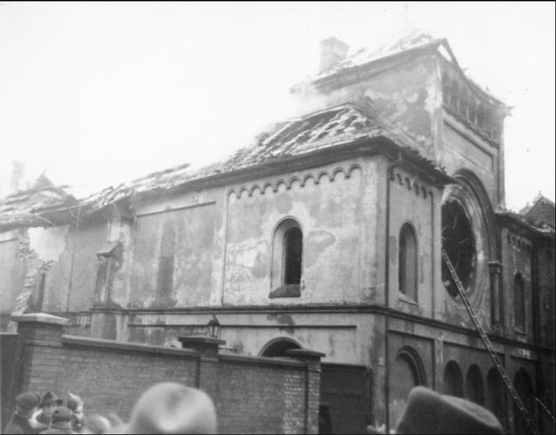 A black and white photograph of a large stone synagogue with a damaged roof and scorched walls, photographed in November 1938 with onlookers gathered outside.